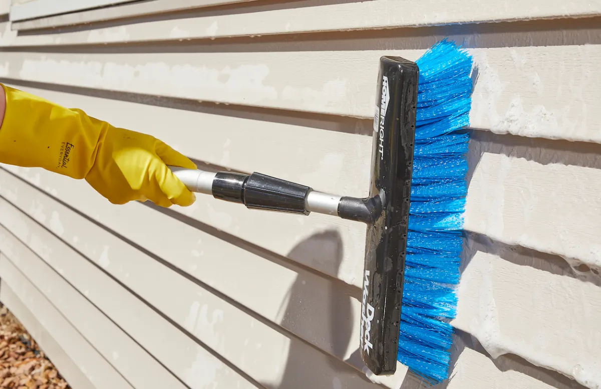 Technician hand-scrubbing vinyl house siding with a soft brush and eco-friendly cleaner to remove dirt and mildew.